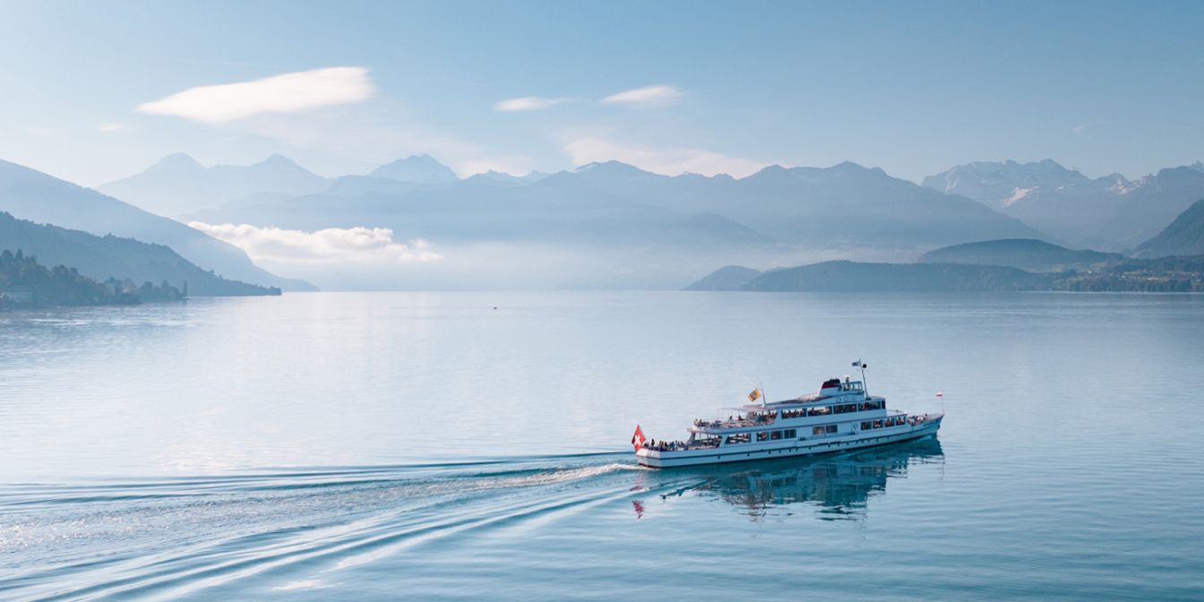 An excursion boat glides leisurely across the calm Lake Thun, while the majestic Alps rise in the background. The gentle morning sun creates a soothing atmosphere and bathes the water and surrounding landscape in delicate shades of blue. ©BLS Schifffahrt AG Boating on Lake Thun