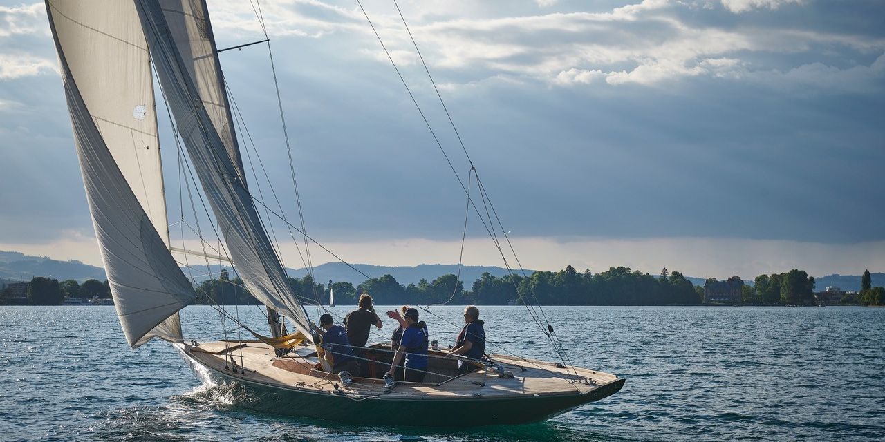Eine Gruppe von Personen segelt auf einem klassischen Segelboot über den Thunersee, umgeben von sanftem Sonnenlicht und malerischer Alpenkulisse. Das perfekte Ambiente für Teamevents, Incentives oder Firmenschulungen direkt am Wasser in der inspirierenden Natur der Region Thun. ©Segelschule Thunersee Segelerlebnis auf dem Thunersee