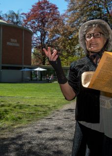 Eine kostümierte Guide steht im Vordergrund auf einem Parkweg und liest aus einem großen Buch, während sie gestikulierend erzählt. Im Hintergrund liegt das Panorama-Rundgebäude im Schadaupark, umgeben von herbstlichen Bäumen – ideal für Teamevents und Rahmenprogramme am Thunersee. ©Thun-Thunersee Tourismus Führung im Schadaupark Thun
