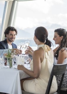 Four business travellers toast each other with white wine in a bright restaurant on Lake Thun, surrounded by a laid table, glasses and flowers. The large windows offer views of the lake and mountains – ideal for company meetings, customer events and networking in the Thun region. Business lunch with lake view