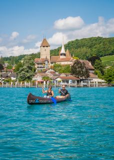 Two people paddle leisurely in a canoe on the turquoise-blue Lake Thun. In the background rises the historic Spiez Castle with its striking church, nestled in a green hilly landscape. The ideal setting for inspiring corporate events or team events by the lake. ©spiez2017_by MikeKaufmann Canoe trip in front of Spiez Castle