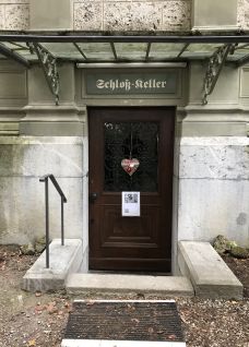 Historischer Zugang zum Schlosskeller: dunkle Holztür mit Dekor, Beschilderung und kleinem Aushang, eingerahmt von Steinmauern.© Schloss Hünegg Eingang zum Schlosskeller mit Holztür und Glasvordach in Thun