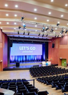 Spacious event auditorium with stage lighting, projection screen and tiered seating—ideal for conferences and presentations near Lake Thun, Switzerland. Auditorium conference hall with stage, large screen and seating in Thun region