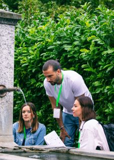 Drei Teilnehmende besprechen Unterlagen am Steinbrunnen im Grünen – informeller Austausch und Teamarbeit in der Region Thun-Thunersee. @FoxTrail Gruppe am Brunnen bei Outdoor-Event in Thun-Thunersee, mit Notizen und Namensbändern