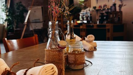 Detailaufnahme eines gedeckten Tisches mit Blumen, Kerzen und Servietten – warme, rustikale Atmosphäre für Business-Events in der Region Thun-Thunersee. ©Frachtraum Thun Stilvoll gedeckter Holztisch mit Vase und Stoffservietten in einer Eventlocation am Thunersee