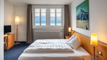 A modern, tranquil double room with a desk, lamps and large windows – the perfect retreat for business guests on Lake Thun. ©Parkhotel, Gunten A bright hotel room with a double bed and views of Lake Thun and the mountains