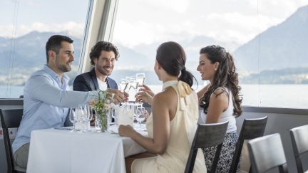 Vier Geschäftsreisende stossen in einem hellen Restaurant am Thunersee mit Weisswein an, umgeben von gedecktem Tisch, Gläsern und Blumen. Durch die grossen Fenster öffnen sich Blick auf See und Berge – ideal für Firmenmeetings, Kundenanlässe und Networking in der Region Thun. ©BLS Schifffahrt Business-Lunch mit Seeblick