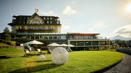 Blick auf ein Seehotel am Thunersee mit moderner Terrasse, Sonnenschirmen und gepflegter Rasenfläche – stilvolle Location für Meetings und Events. ©Strandhotel Belvédère Seehotel mit Terrasse und Eventrasen am Thunersee bei Abendsonne
