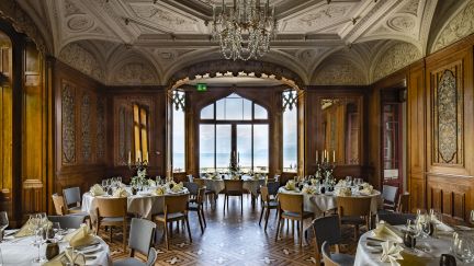 Stilvoller Eventraum mit Kronleuchter, Holzvertäfelung und festlich gedeckten Tischen; durch die Fenster öffnet sich der Blick auf den Thunersee. ©Schloss Schadau Eleganter Bankettsaal mit gedeckten Tischen und Seeblick in der Thun-Thunersee Region