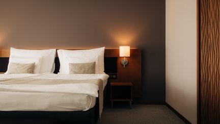 Gemütliches Doppelzimmer in einem Hotel am Thunersee mit sanften Lichtelementen und schicker Einrichtung. Helle Bettwäsche und elegante Wandverkleidungen sorgen für eine entspannte Wohlfühlatmosphäre – ideal für einen erholsamen Incentive Aufenthalt in Thun, Schweiz. ©Deltapark Modernes Doppelzimmer mit Ambiente