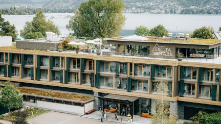 Moderne Aussenansicht des Delta Park Hotels in Thun, eingebettet in eine grüne Umgebung mit direktem Blick auf den Thunersee. Die großzügige Terrasse und die stilvolle Architektur laden zum Verweilen ein. Im Hintergrund sind die Alpen und das Wasser zu erkennen. Aussenansicht Delta Park Hotel am Thunersee