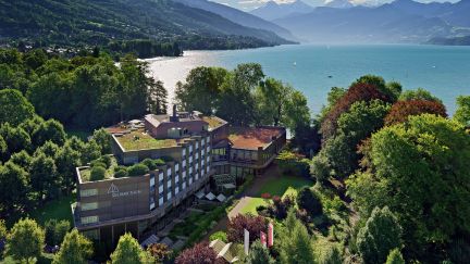 Luftaufnahme eines Seminarhotels am Ufer des Thunersees, umgeben von Wald und Gärten – ruhige Lage mit Blick auf See und Berge. ©Seepark Panoramablick auf ein Seminarhotel am Thunersee mit Alpenkulisse