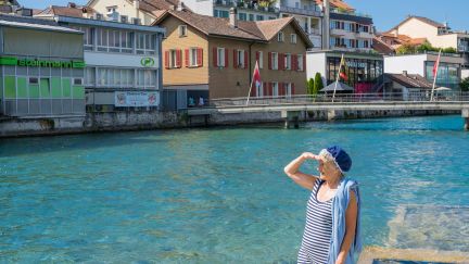 Sommerliche Szene am klaren Wasser: Spaziergängerin am Steg, dahinter Hafenbecken und historische Häuserzeile in Thun am Thunersee. Themenführungen sind ideal als Rahmenprogramm nach einem langen Seminartag. ©Thun-Thunersee Tourismus Frau am Ufer mit Blick auf Hafen und Altstadthäuser in Thun am Thunersee