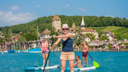 Three people enjoy stand-up paddleboarding on Lake Thun in bright sunshine. In the background, the picturesque Spiez Castle rises against an idyllic Alpine backdrop. Ideal for team events and corporate experiences in an inspiring lakeside setting. ©spiez2017_by MikeKaufmann Stand-up paddleboarding in front of Spiez Castle