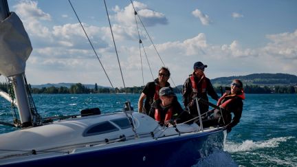 Vier Personen steuern gemeinsam ein Segelboot bei frischem Wind auf dem Thunersee. Die Teilnehmer tragen Schwimmwesten und konzentrieren sich auf das Zusammenspiel an Bord. Im Hintergrund sind die Voralpen und ein klarer Himmel zu sehen – ideale Bedingungen für Teambuilding-Events oder Firmenausflüge am Wasser. @Segelschule Thunersee Teamsegeln auf dem Thunersee