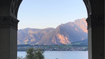 A stunning view through a historic archway of Lake Thun and the mountains in the soft morning light. The tranquil lakeside setting, with a well-tended garden in the foreground, creates an inspiring atmosphere – ideal for conferences and corporate events by the lake in the Thun region. Panoramic view of Lake Thun