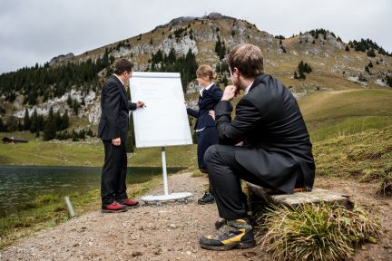 Three businesspeople in suits hold a creative meeting outdoors by a lake with impressive Alpine scenery. A flipchart serves as the central working tool. The natural environment promotes inspiring ideas and undisturbed exchange, ideal for company workshops and team building on Lake Thun. ©Stockhorn Creative outdoor meeting in the Alps