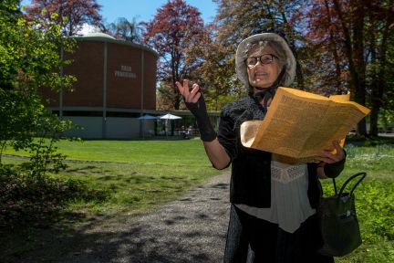 A costumed guide stands in the foreground on a park path and reads from a large book while gesturing as she tells her story. In the background is the panoramic rotunda in Schadau Park, surrounded by autumnal trees – ideal for team events and supporting programmes on Lake Thun. Guided tour of Schadau Park, Thun