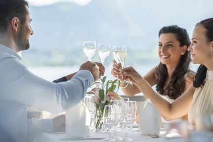 Drei Personen erheben Weingläser beim stilvollen Dinner am See – helles Ambiente, Alpenpanorama und ideal für MICE-Events in der Region Thun. ©BLS-Schifffahrt Business-Gäste stossen beim Dinner am Thunersee mit Blick auf die Alpen an