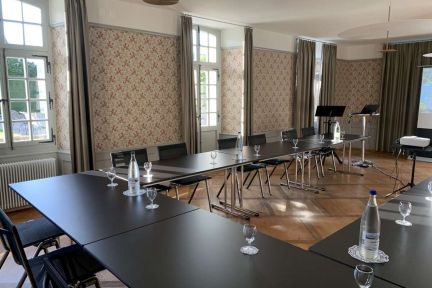 Bright seminar room with U-shaped tables, water glasses and carafes – ideal for meetings, workshops or training courses. High windows, classic wallpaper and parquet flooring create a calm, professional atmosphere on Lake Thun in the canton of Bern, perfect for companies and associations. ©Spiez Castle Seminar room in the U-setting