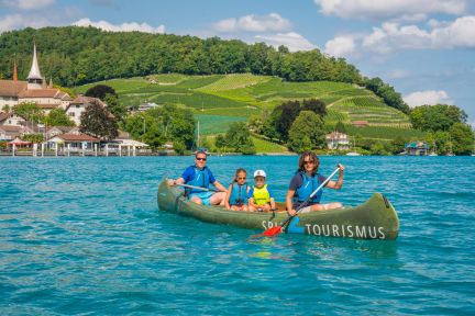 A group of motivated participants paddle together in a canoe on the turquoise-blue Lake Thun. The picturesque vineyards of Spiez and the church can be seen in the background. An ideal team-building experience for companies and organisations in an inspiring natural setting with a magnificent Alpine panorama. ©spiez2017_by MikeKaufmann Team experience: canoe trip on Lake Thun