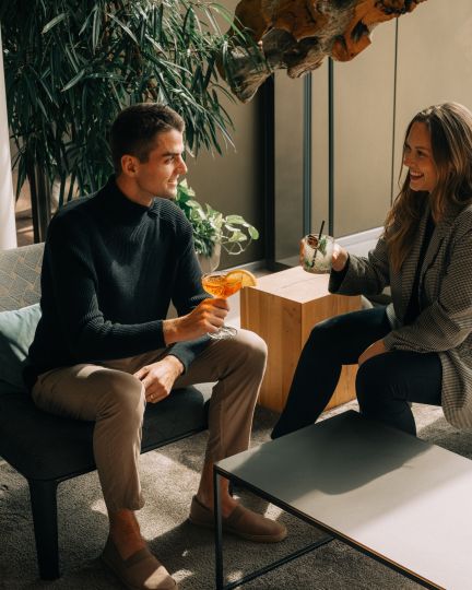 Zwei Gäste genießen entspannt ihre Getränke in einer stilvoll eingerichteten Hotellobby in Thun. Natürliche Lichtquellen, moderne Möbel und eine gemütliche Atmosphäre schaffen einen perfekten Ort für angenehme Gespräche. Die freundliche Stimmung unterstreicht das hochwertige Hotelerlebnis am Thunersee. Entspannter Drink in moderner Hotellobby