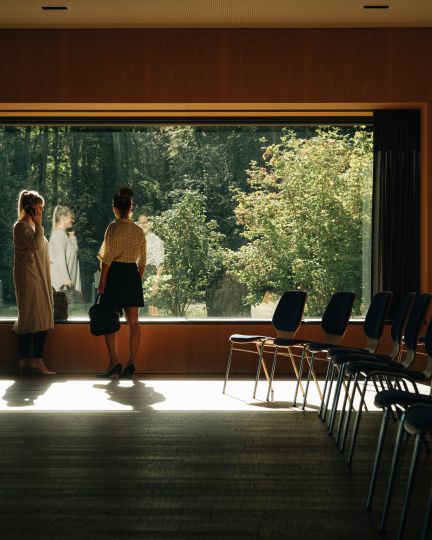A bright seminar room with large windows offering views of a green, peaceful forest. Two people chat relaxed in the light shining through the window, while rows of empty chairs invite visitors to take a seat. The scene conveys an open, inspiring atmosphere for creative meetings surrounded by nature near Thun. ©Deltapark Modern seminar room with forest view