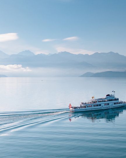 An excursion boat glides leisurely across the calm Lake Thun, while the majestic Alps rise in the background. The gentle morning sun creates a soothing atmosphere and bathes the water and surrounding landscape in delicate shades of blue. ©BLS Schifffahrt. Boating on Lake Thun
