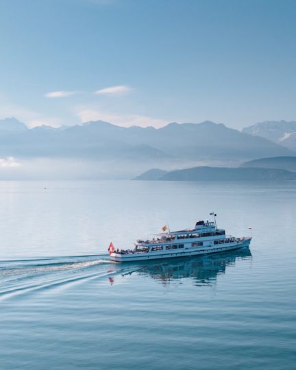 Ein Ausflugsschiff gleitet gemächlich über den ruhigen Thunersee, während sich im Hintergrund majestätische Alpen erheben. Die sanfte Morgensonne sorgt für eine beruhigende Atmosphäre und lässt das Wasser und die umliegende Landschaft in zarten Blautönen leuchten. ©BLS Schifffahrt. Schifffahrt auf dem Thunersee