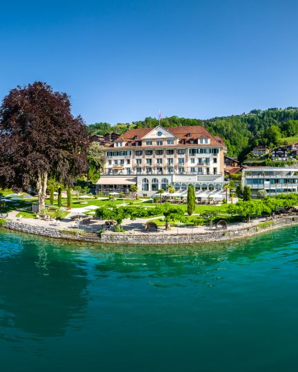 Panoramablick auf ein Seehotel am Ufer des Thunersees – gepflegte Gartenanlage, Bootshaus und ruhige Wasserfläche für stilvolle Business-Events. ©Parkhotel Gunten Hotel am Thunersee mit Seeterrasse und Park, umgeben von Alpenpanorama