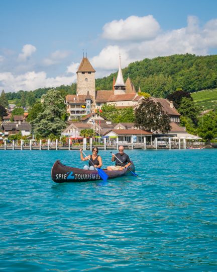 Zwei Personen paddeln entspannt in einem Kanu auf dem türkisblauen Thunersee. Im Hintergrund erhebt sich das historische Schloss Spiez mit seiner markanten Kirche, eingebettet in grüne Hügellandschaft. Ideale Szenerie für inspirierende Firmenanlässe oder Teamevents am See. ©spiez2017_by MikeKaufmann Kanufahrt vor Schloss Spiez