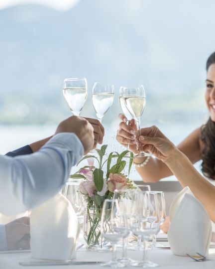 Drei Personen erheben Weingläser beim stilvollen Dinner am See – helles Ambiente, Alpenpanorama und ideal für MICE-Events in der Region Thun. ©BLS-Schifffahrt Business-Gäste stossen beim Dinner am Thunersee mit Blick auf die Alpen an