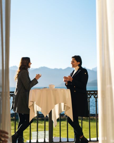 Zwei Geschäftsfrauen genießen eine entspannte Kaffeepause an einem runden Stehtisch auf einer sonnigen Terrasse mit freiem Blick auf den funkelnden Thunersee und die Alpen. Ideal für inspirierende Gespräche und Networking in stilvoller Umgebung. ©Thun-Thunersee Tourismus Business-Kaffeepause mit Blick auf den Thunersee
