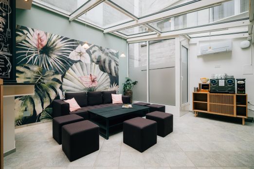 Moderner Loungebereich für Business-Events: Sofa, Poufs und Couchtisch unter Glasdach, mit grossflächigem Pflanzenmotiv an der Wand. ©Hotel Krone Helle Event-Lounge mit Glasdach, Sitzgruppe und Tropenwandbild