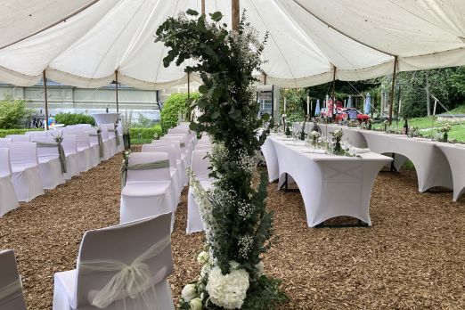 Ein festlich geschmücktes Zelt mit weißen Stuhlhussen und elegant dekorierten Tischen auf naturbelassenem Boden. Frische Blumenarrangements und viel Grün sorgen für eine inspirierende Atmosphäre – ideal für Firmenveranstaltungen und Events am Thunersee im Kanton Bern. © Schloss Hünegg Stilvoll dekoriertes Eventzelt am Thunersee