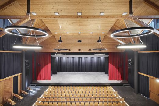 Blick von hinten in einen Holzsaal mit Bühne, roten Vorhängen und Reihenbestuhlung – ideal für Tagungen und Firmenanlässe in der Thunersee-Region. ©Gemeinde Spiez Moderner Veranstaltungssaal mit Bühne, Reihenbestuhlung und Ringlichtern in Thun