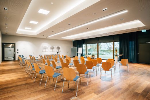 Heller Seminarraum mit großen Fenstern und stilvoller Holzoptik, ausgestattet mit bequemen Stühlen in Reihenanordnung. Modernes Ambiente, freundliche Atmosphäre und Zugang zur Terrasse schaffen ideale Bedingungen für erfolgreiche Workshops und Veranstaltungen in Thun am Thunersee. Moderner Seminarraum mit Tageslicht
