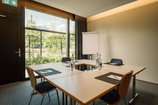 Heller Seminarraum mit großen Fenstern und Blick ins Grüne, ausgestattet mit Flipchart, Notizmaterial und Mineralwasser. Die freundliche Atmosphäre und die ruhige Umgebung bieten optimale Voraussetzungen für erfolgreiche Meetings in Thun am Thunersee. Moderner Seminarraum mit Tageslicht
