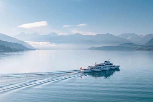 Ein Ausflugsschiff gleitet gemächlich über den ruhigen Thunersee, während sich im Hintergrund majestätische Alpen erheben. Die sanfte Morgensonne sorgt für eine beruhigende Atmosphäre und lässt das Wasser und die umliegende Landschaft in zarten Blautönen leuchten. ©BLS Schifffahrt. Schifffahrt auf dem Thunersee