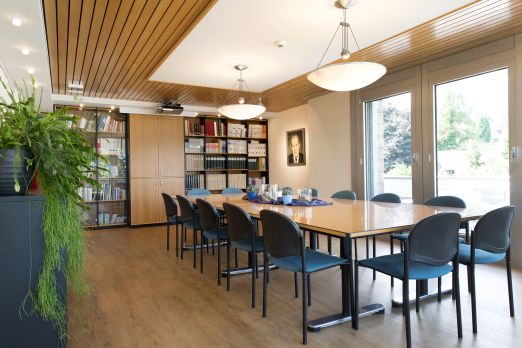 Moderner und einladender Besprechungsraum mit großen Fenstern, hellen Holzelementen und komfortabler Bestuhlung. Der Raum eignet sich optimal für Meetings, Workshops oder Seminare kleiner und mittlerer Unternehmen in der Region Thunersee. Pflanzen und Bücherregale schaffen eine angenehme Arbeitsatmosphäre. ©Seaside Spiez Heller Tagungsraum mit natürlichem Licht