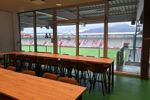 Modernes Meeting-Setting mit Holztischen und Reihenbestuhlung; durch die Glasfront fällt der Blick direkt auf das Spielfeld im Stadion Thun. ©Stockhorn Arena Seminarraum mit Panoramafenster und Blick ins Stadion Thun