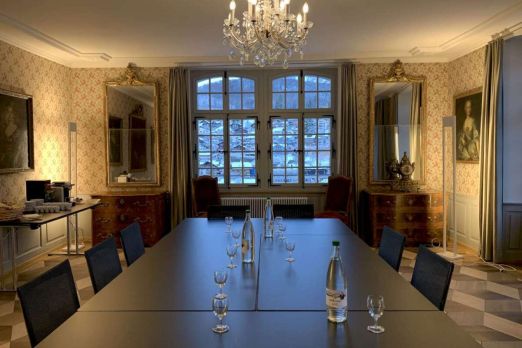 Stilvoller Sitzungsraum für Business-Meetings: gedeckte Tafel mit Wasserflaschen, Kronleuchter und grosse Fensterfront im Abendlicht. ©Schloss Spiez Eleganter Meetingraum mit langer Tafel, Kronleuchter und Blick auf Thun