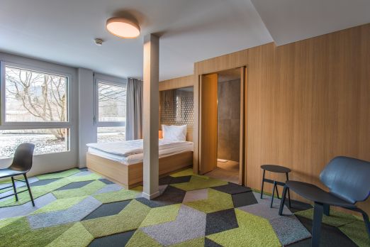 Helles Doppelzimmer mit geometrischem Teppich, Sitzbereich und Badzugang – ideal für Businessgäste und Eventteilnehmende in der Region Thunersee. ©The Lab Hotel Modernes Hotelzimmer mit Doppelbett, Holzverkleidung und großem Fenster in Thun