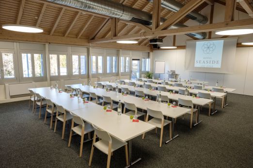 Heller Tagungsraum mit Holzdecke, U-Bestuhlung, Präsentationsleinwand und viel Tageslicht – ideal für Seminare und Workshops am Thunersee. ©Seaside Spiez Seminarraum mit U-Bestuhlung und Beamer in der Region Thun-Thunersee