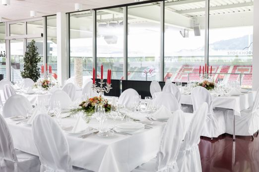 Weiss eingedeckte Tische mit Blumen und roten Kerzen in einem lichtdurchfluteten Saal – stilvoller Rahmen für Business-Events am Thunersee. ©Stockhorn Arena Festlich gedeckte Banketttische in heller Eventlocation mit Panoramafenstern