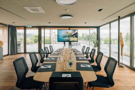 Lichtdurchfluteter Seminarraum mit großflächigen Fenstern, modernem Mobiliar und Aussicht ins Grüne. Der große Konferenztisch ist stilvoll gedeckt, mit Getränken, Schreibmaterial und Präsentationsbildschirm. Ideal für Meetings und Workshops in inspirierender Umgebung am Thunersee. Moderner Seminarraum mit Panoramablick