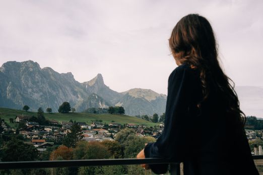 Eine Frau genießt die malerische Aussicht von einem Hotelbalkon auf ein idyllisches Schweizer Dorf und die imposanten Berge der Berner Alpen. Die entspannte Atmosphäre und das weite Panorama laden zum Verweilen ein und machen den Aufenthalt in Thun besonders. ©Deltapark Aussicht vom Hotelbalkon auf die Berner Alpen
