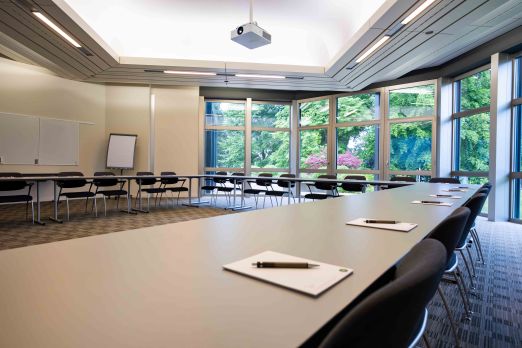 Moderner, lichtdurchfluteter Konferenzraum mit großen Fenstern und Blick ins Grüne. Hufeisenförmige Tischanordnung, Stühle, Whiteboard und Flipchart schaffen das perfekte Umfeld für Meetings, Seminare oder Workshops in inspirierender Atmosphäre am Thunersee. Heller Konferenzraum mit Gartenblick - Basel