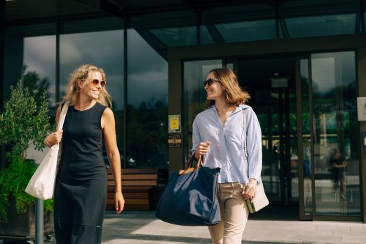 Zwei lächelnde Frauen mit Taschen verlassen gemeinsam einen modernen Hoteleingang in Thun. Die Glasfassade des Gebäudes spiegelt die Umgebung wider, während warme Sonnenstrahlen für eine einladende Stimmung sorgen. Das Bild vermittelt Gastfreundschaft und einen gelungenen Start in den Aufenthalt am Thunersee. ©Deltapark Freundlicher Empfang am Hoteleingang