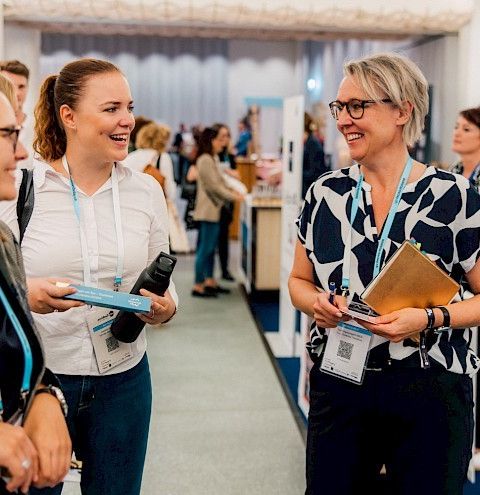 Drei Berufsfrauen unterhalten sich angeregt in einer modernen Kongressumgebung. Im Hintergrund sind weitere Teilnehmer und Messestände zu sehen. Diese Szene spiegelt die offene und inspirierende Atmosphäre für Geschäftstreffen und Events in der Region Thunersee wider. ©Assistants Day Zürich Persönliche Beratung von Thun-Thunersee Tourismus
